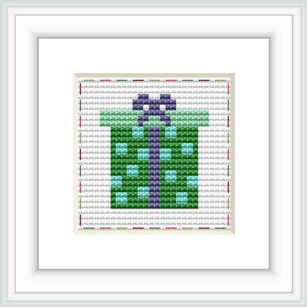 This is a framed cross stitch image depicting a stylized wrapped gift with a bow, primarily in green and purple hues. It's set on a white background with red and grey decorative stitches around the border.