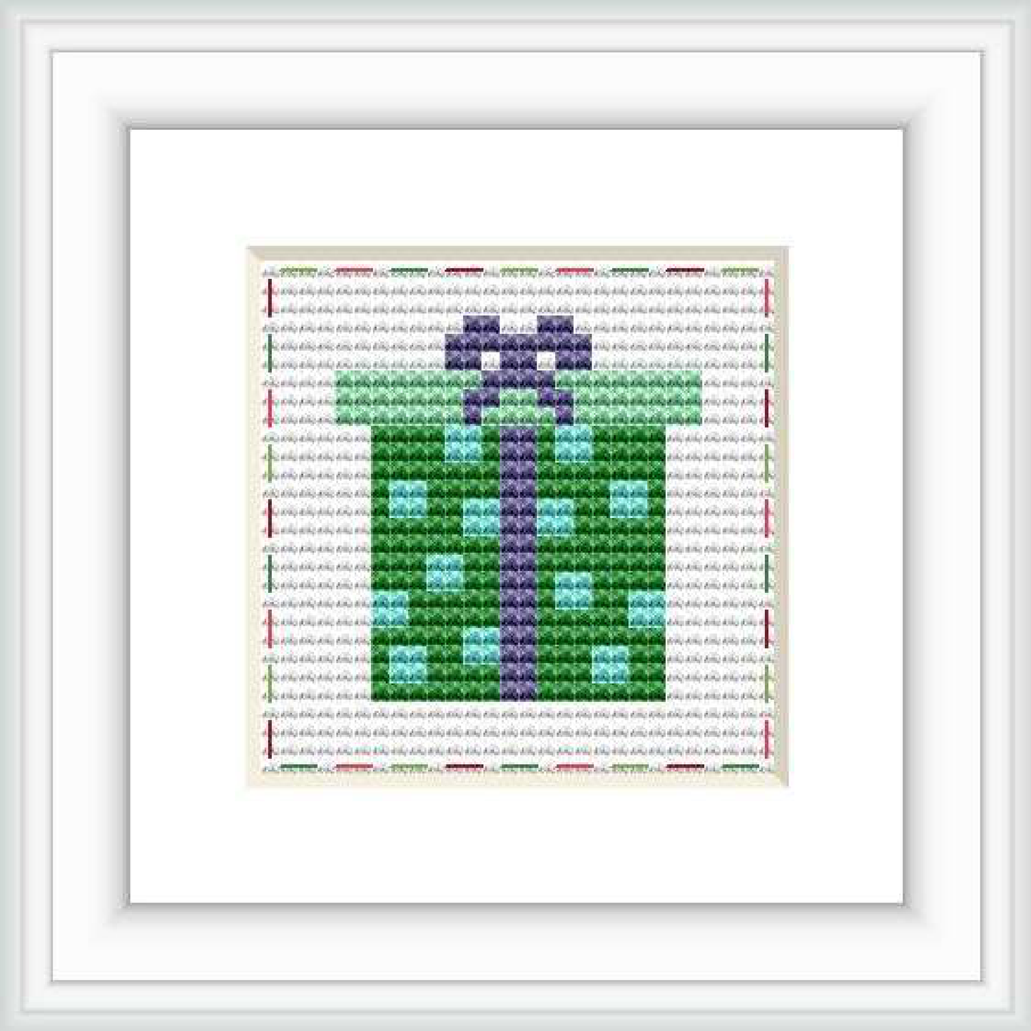 This is a framed cross stitch image depicting a stylized wrapped gift with a bow, primarily in green and purple hues. It's set on a white background with red and grey decorative stitches around the border.