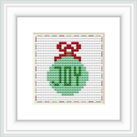 The image features a framed cross stitch of a green Christmas ornament with a red ribbon on a patterned light grey background. The cross stitch is centralized within a white mat and a simple black frame.