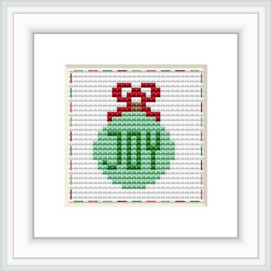 The image features a framed cross stitch of a green Christmas ornament with a red ribbon on a patterned light grey background. The cross stitch is centralized within a white mat and a simple black frame.