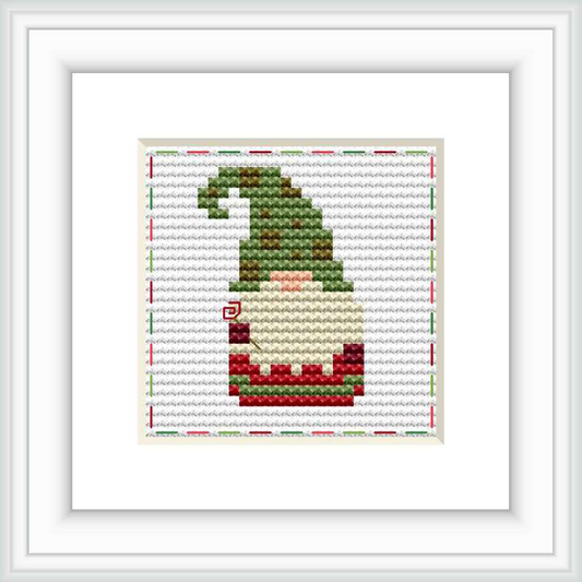 A framed cross stitch pattern depicting a stylized gnome in profile, wearing a green hat, with a stitched white border on a fabric background.