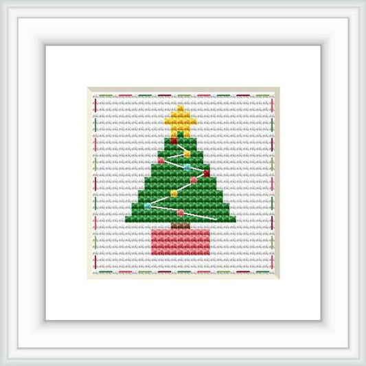 The image depicts a framed cross stitch pattern featuring a Christmas tree with a yellow star and multicolored ornaments, set against a white background with a grey grid. A dark red ribbon is woven through the branches, and the tree stands on a pink base. The cross stitch is surrounded by a white border.