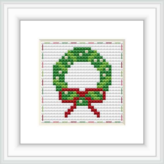 The image shows a framed cross stitch pattern of a holiday wreath with a red bow, against a white background. The stitches are visible and display a 28 by 28 grid pattern.