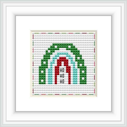 The image displays a framed cross stitch design with a central rainbow motif along with the words 'Ho Ho Ho' in the center. It is stitched on a white fabric with a grid of colorful threads forming the rainbow and letters.