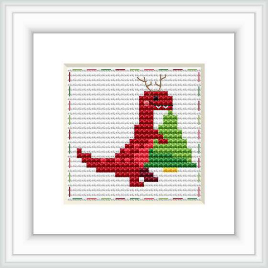 This image showcases a framed cross stitch pattern featuring a stylized red and green dinosaur against a white background with grey grid lines. The framed piece is displayed against a plain background.