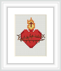 The image showcases a cross stitch pattern framed and displayed on a wall. The pattern features a stylized red heart with a yellow and orange flame on top, all outlined by brown thorny branches, on a white stitched background.