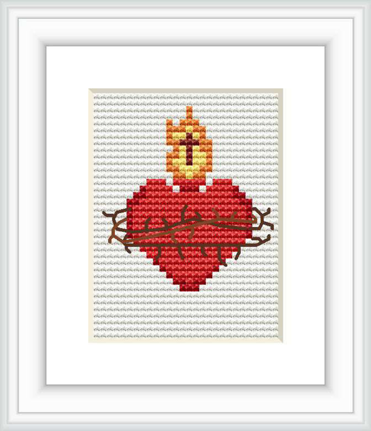 The image showcases a cross stitch pattern framed and displayed on a wall. The pattern features a stylized red heart with a yellow and orange flame on top, all outlined by brown thorny branches, on a white stitched background.