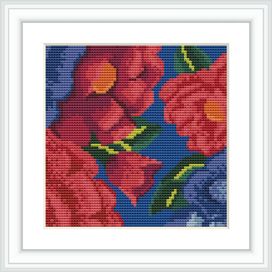 The image displays a framed cross stitch pattern of red and blue flowers with green leaves set against a dark blue background, placed on a light surface.