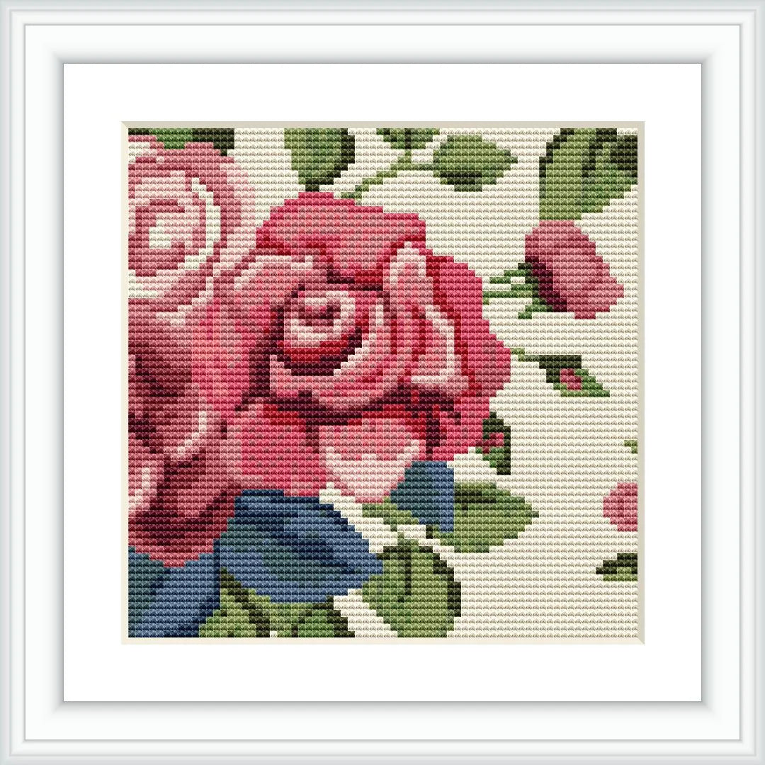 The image depicts a framed cross stitch pattern of roses. There are prominent large red roses with smaller flowers and foliage against an off-white background. The stitching is detailed, neatly showcasing the various shades and colors.