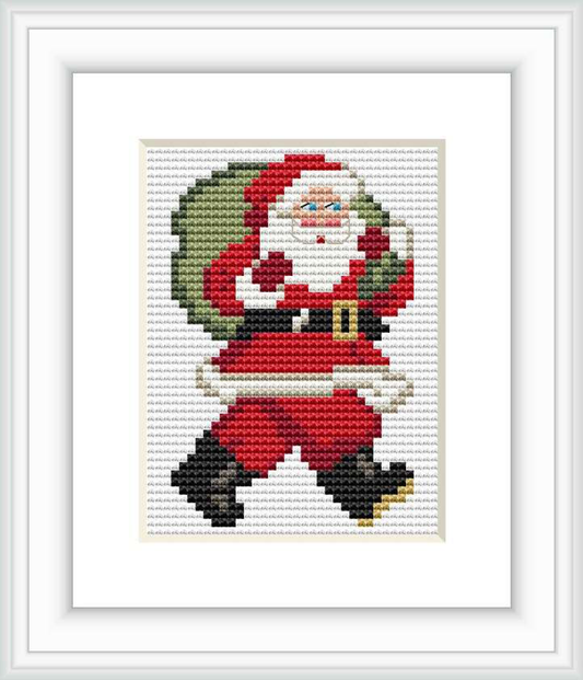 The image depicts a framed cross stitch pattern of Santa Claus standing with a sack over his shoulder. Santa is wearing his classic red and white outfit with black boots and a belt, against a plain white background.