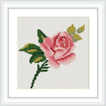 The image features a framed cross stitch pattern of a pink rose with green leaves and yellow accents on a white background.