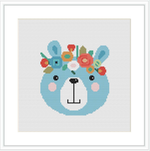 The image displays a cross stitch pattern of a stylized bear face in the center with a variety of flowers adorning its head. The bear has a light blue face, blushed cheeks, and a simple yet cute expression. The background is white, emphasizing the pattern's colors.