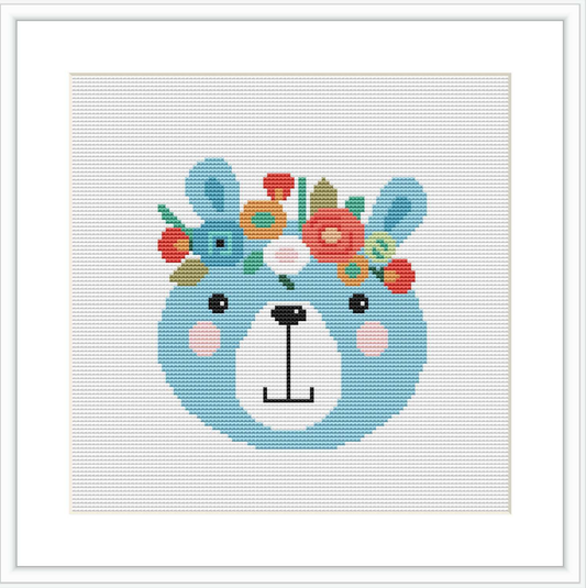 The image displays a cross stitch pattern of a stylized bear face in the center with a variety of flowers adorning its head. The bear has a light blue face, blushed cheeks, and a simple yet cute expression. The background is white, emphasizing the pattern's colors.