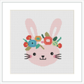 The image depicts a cross stitch pattern showing a stylized pink bunny with flowers and leaves on its head. The bunny has a cheerful expression with its eyes closed and mouth in a content smile. The embroidery is set within a square grid, framed by a simple white border.
