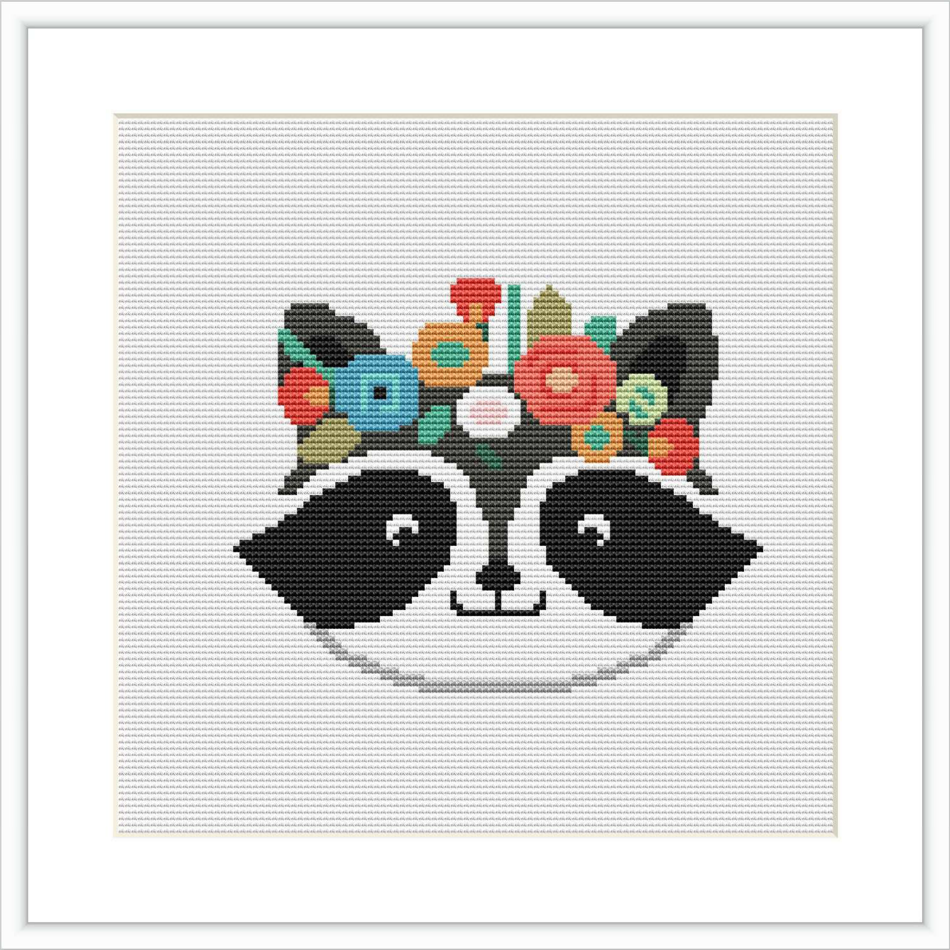 The image showcases a cross stitch pattern of a stylized raccoon's face with eyes, nose, and ears in black and white. Above the eyes, there is a colorful collection of stitched flowers in various colors like pink, green, and blue, representing a floral crown.