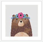 A brown bear with a floral crown on its head, comprising pink and blue flowers. The pattern is square with a small white border framing the design on a grid-background indicative of cross stitch fabric.