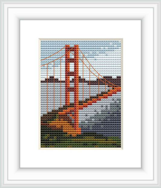 The image is a framed cross stitch depiction of the Golden Gate Bridge in San Francisco. The composition includes the bridge in the foreground with supporting cables and towers, and stylized clouds in the sky positioned above.