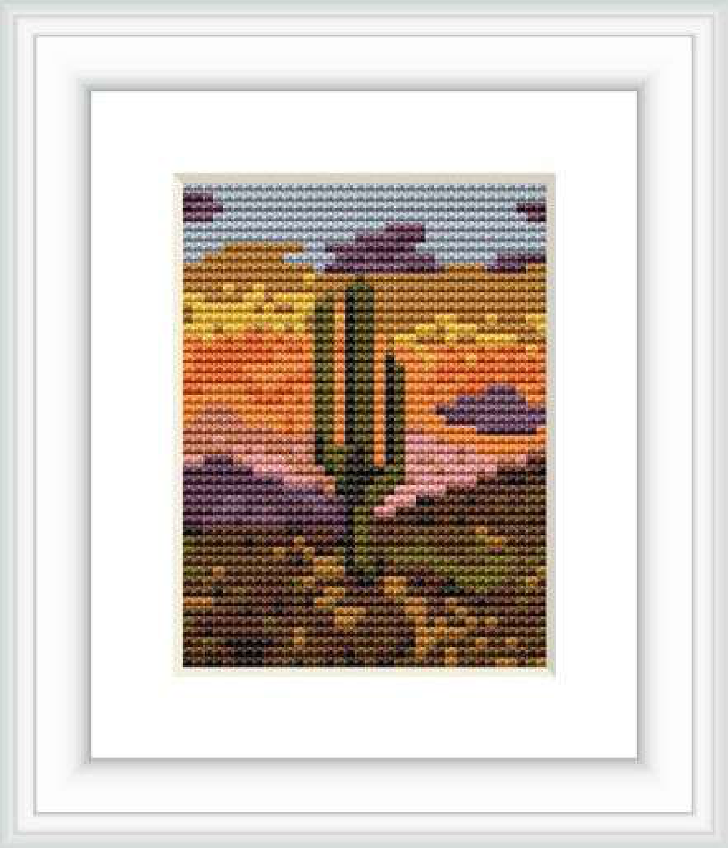 The image depicts a cross stitch embroidery of a desert landscape at dusk. There's a prominent cactus in the center with a multicolored sky made up of yellow, orange, purple, and blue hues in the background.