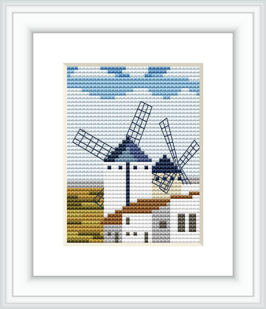 The image depicts a cross-stitch pattern featuring a white windmill with blue accents and spinning blades, set against a backdrop of a light blue sky with clouds. Below, there is a representation of brown and green fields, alongside a white building with a brown roof, indicative of a rural Spanish landscape.