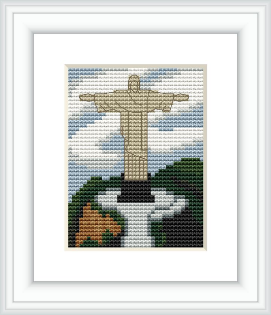 The image displays a cross stitch pattern of the Christ the Redeemer statue with a backdrop of blue sky and green foliage. The statue is depicted in white and beige, standing tall with arms outstretched, atop a black base against the foliage.