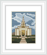 The image shows a framed cross stitch pattern depicting a white temple-like building with a tall spire, surrounded by green bushes, with a backdrop of a blue sky interspersed with clouds.