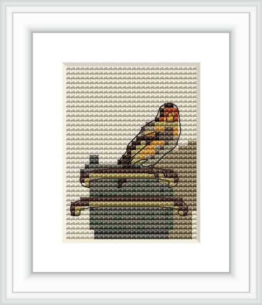 The image displays a cross stitch pattern of a goldfinch perched on top of a feeder, with a mostly muted color palette and a realistic depiction.