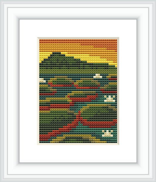 The image depicts a cross stitch pattern framed in white, showcasing a stylized mountain landscape with varying shades of greens, browns, yellows, and hints of red.