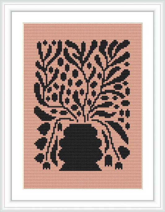 The image depicts a cross stitch pattern of stylized black flowers on a pink background, framed in a simple white frame.