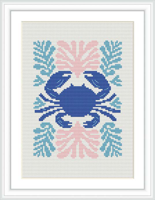 The image features a framed cross stitch pattern of a stylized crab centered on a white fabric. Surrounding the crab are coral-like embellishments in various shades of blue, pink, and coral. The pattern is rendered with a variety of stitches to depict the distinct form of the crab and the decorative elements.