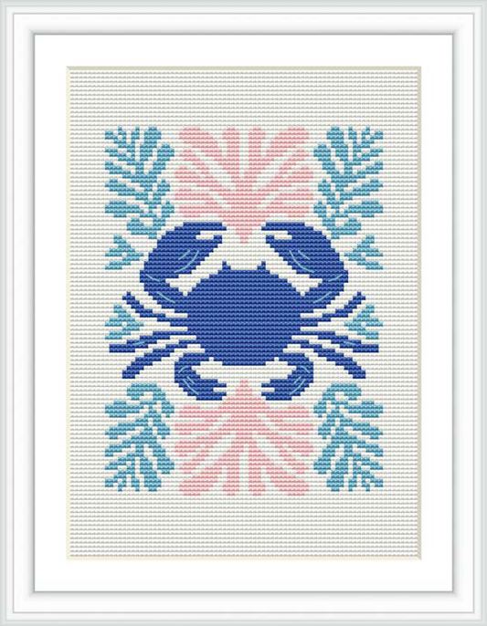 The image features a framed cross stitch pattern of a stylized crab centered on a white fabric. Surrounding the crab are coral-like embellishments in various shades of blue, pink, and coral. The pattern is rendered with a variety of stitches to depict the distinct form of the crab and the decorative elements.