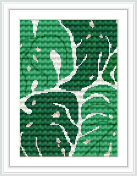 The image shows a framed cross stitch pattern resembling Monstera plant leaves in various shades of green, with the pattern visible against a white background.