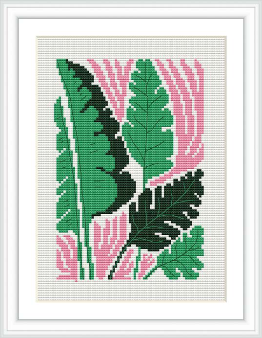 The image depicts a framed cross stitch pattern that features multiple monstera plant leaves in various shades of green with pink accents, stitched onto a white fabric. The frame is also white, and the pattern is viewed from the front showcasing the completed design.