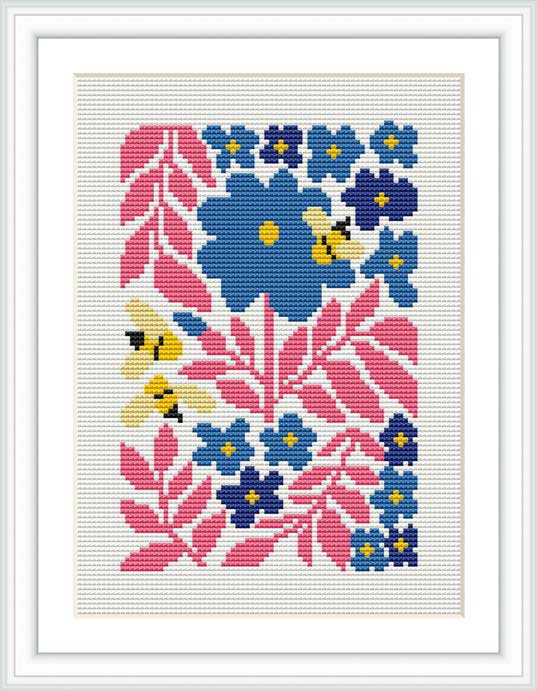 The image shows a framed cross stitch design featuring a pattern of blue and pink flowers with green leaves. It is displayed against a white fabric background, and the pattern is centralized within the frame.