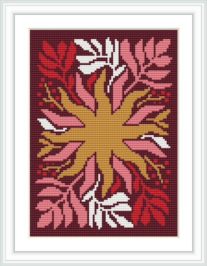 The cross stitch pattern showcases a central symmetrical floral design with petals in shades of red and pink, accented by white and gold. It is framed within a white mat and dark frame, set against a contrasting burgundy background.