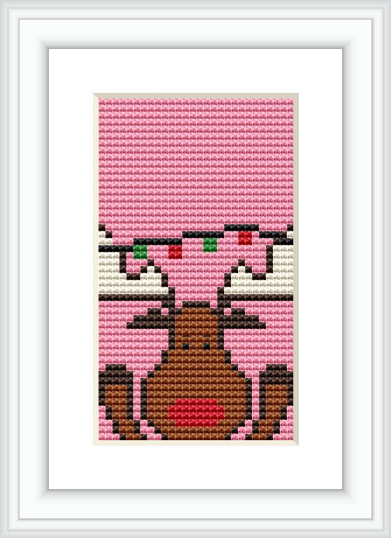 The image displays a cross stitch pattern within a frame. The design depicts a stylized Rudolph the Red-Nosed Reindeer with a red nose and antlers, set against a pinkish background.