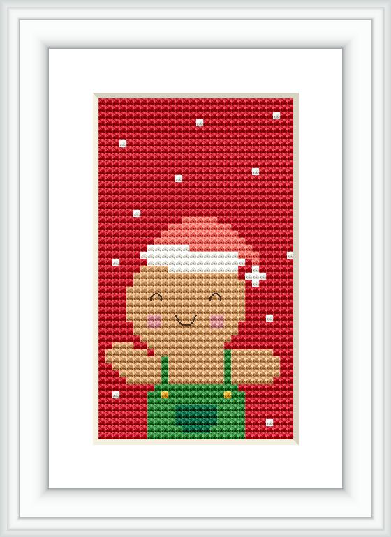 The image shows a framed cross stitch pattern on a wall. The design is centered on a gingerbread figure with a white squiggly line on its head, resembling icing, and a green bib apron. The background is predominantly red with sporadic white dots that simulate falling snow or sugar sprinkles.