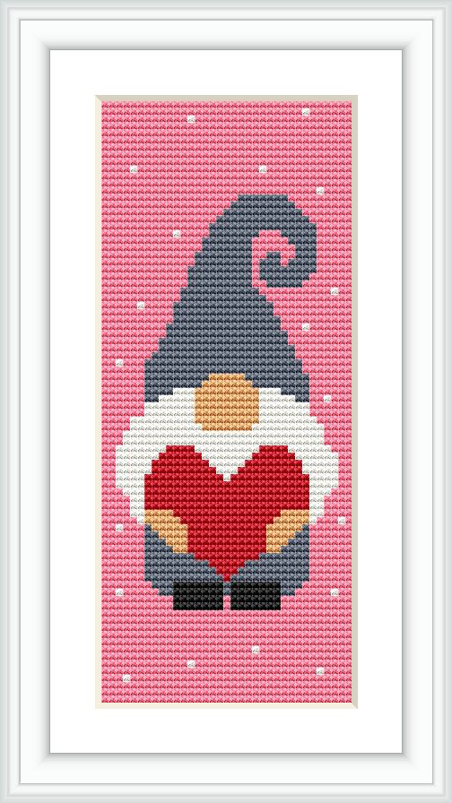 A framed cross stitch pattern of a stylized Santa figure against a pink background with white snowflake accents. The Santa wears a large hat and holds a heart.