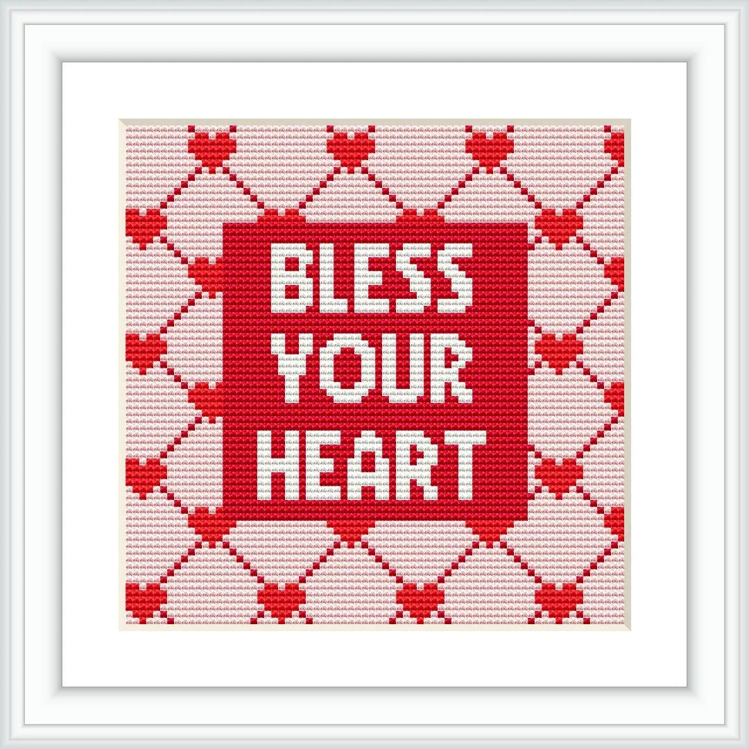 The image shows a cross stitch design framed in white, featuring the phrase 'BLESS YOUR HEART' centered on a red background, with a pattern of white hearts and diamonds decorating the border area.